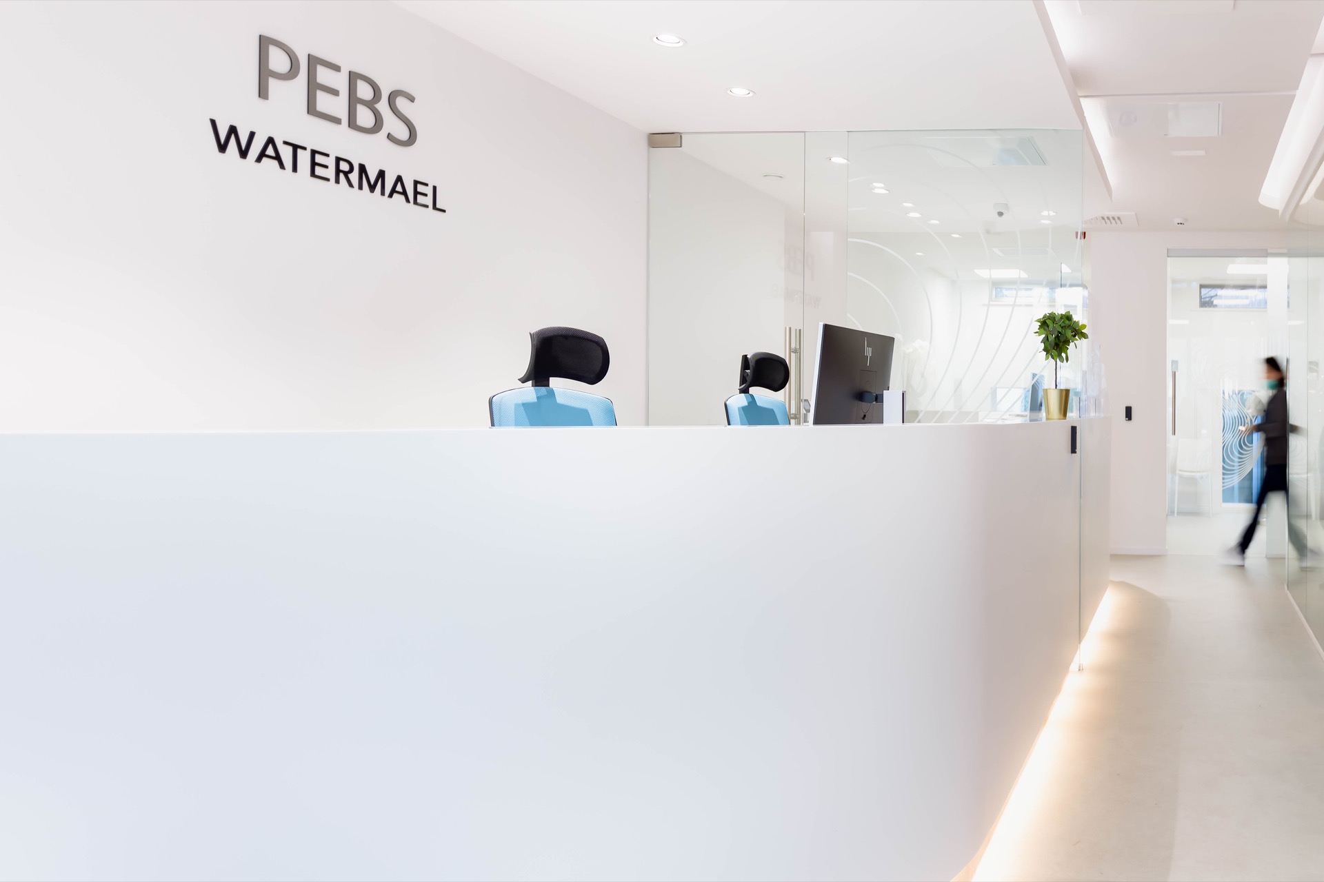 PEBS Watermael — comptoir d'accueil sculpté blanc avec logo en relief, perspective enfilade vers le couloir lumineux