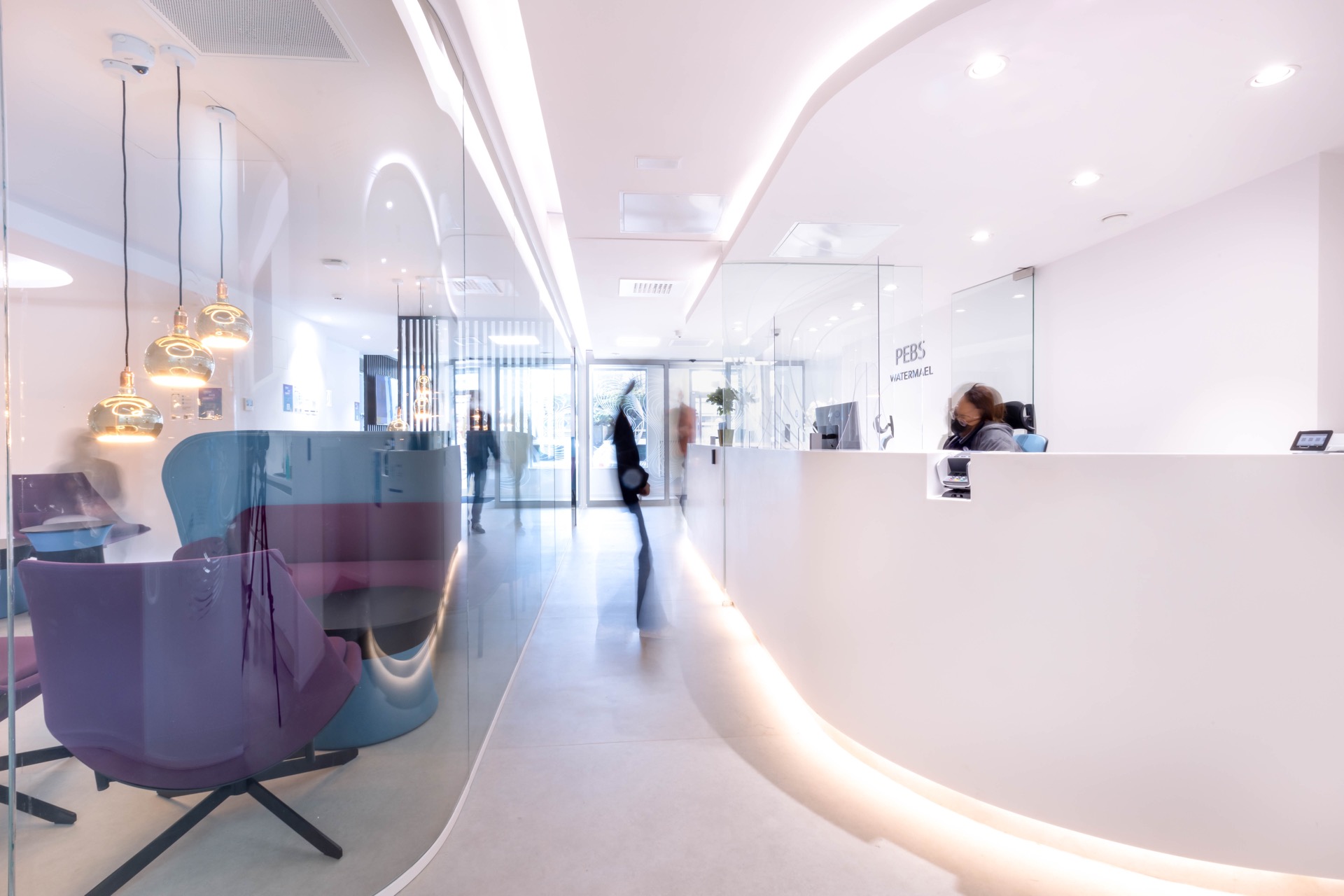 PEBS Watermael — comptoir d'accueil sculpté blanc avec logo en relief, perspective enfilade vers le couloir lumineux
