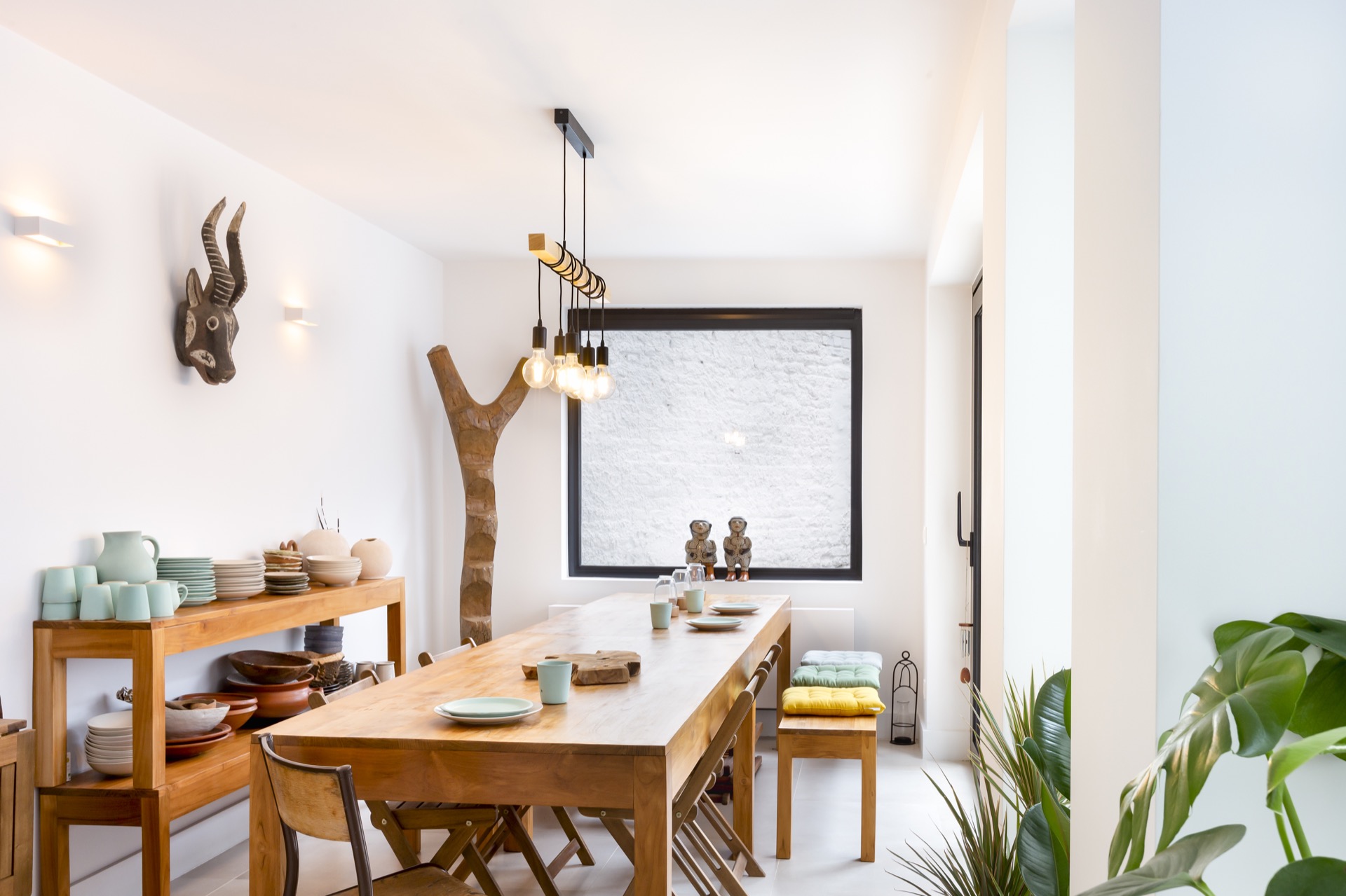 Vandenbusch — salle à manger ethno-chic, suspension Edison ligne sur tronc d'arbre brut, vaisselier teck et céramique céladon, tête d'antilope sculptée